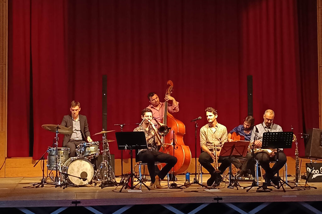 Photo montrant le groupe Woo Katz au festival Vichy Jazzy, le dimanche 20 octobre à 17 h, à la salle des fêtes de Vichy. On y voit 5 hommes assis jouant de leur instrument et un homme debout jouant également de son instrument. Les six artistes jouent du Jazz New Orleans sur scène, face à un public assis.