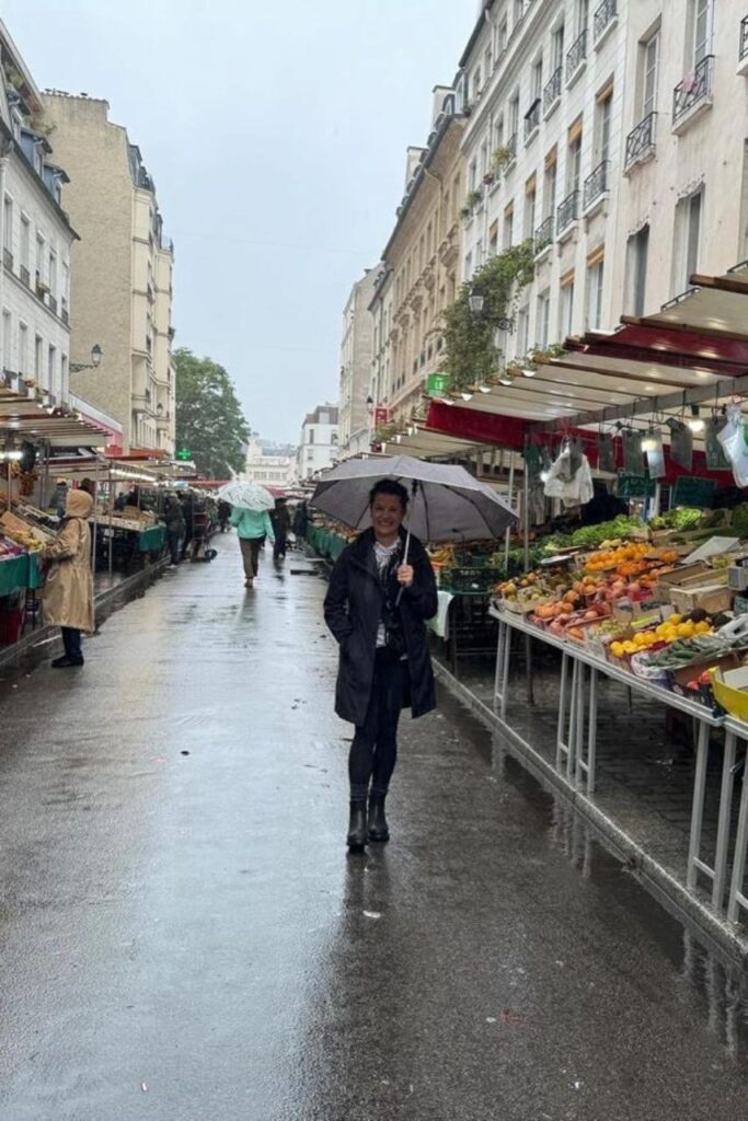 Le marché de Moulins un jour de pluie. Les passants tous couverts, choisissent leurs produits. Au premier plan, une femme qui marche sourit sur la photo.