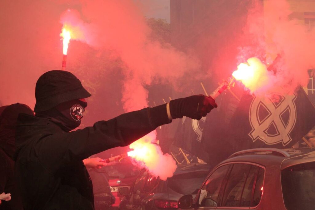 Un militant habillé en noir, à visage couvert, brandit des fumigènes de fumée rouge dans une rue parisienne.