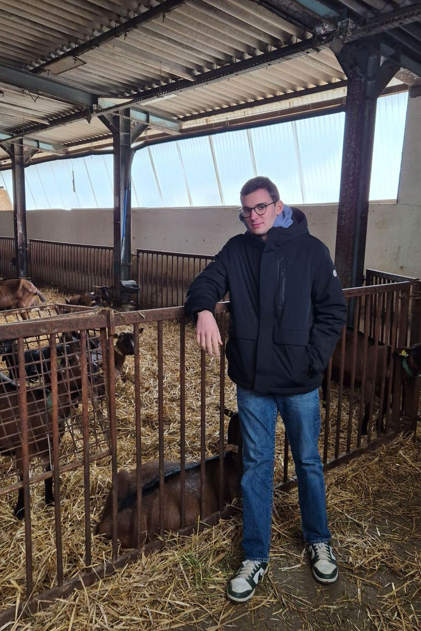 : Matthias Boyé accoudé à la clôture d’un enclos, dans le hangar de la chèvrerie du Lycée de Ressins. Il se tient debout, une main dans la poche de sa veste bleue. Une dizaine de chèvres se trouvent derrière lui dans l’enclot, dont une qui sort sa tête des barrières.